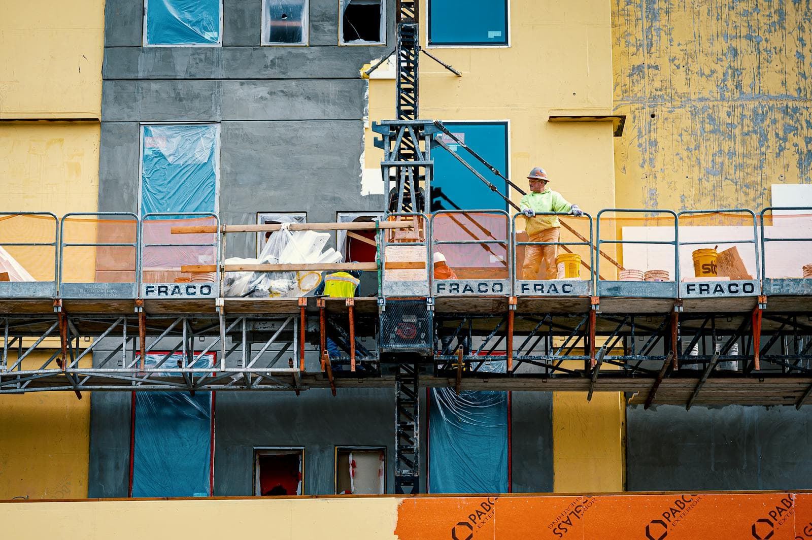 Bygningsarbeider i hjelm og gul jakke står på en heisbar arbeidsplattform mot en bygningsfasade.