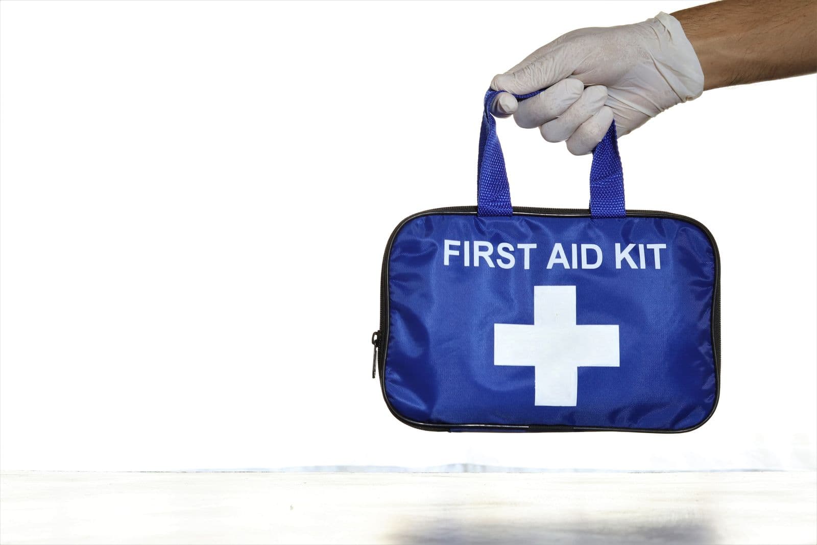 En hånd med hvit hanske holder en blå førstehjelpsveske merket FIRST AID KIT med hvitt kors.