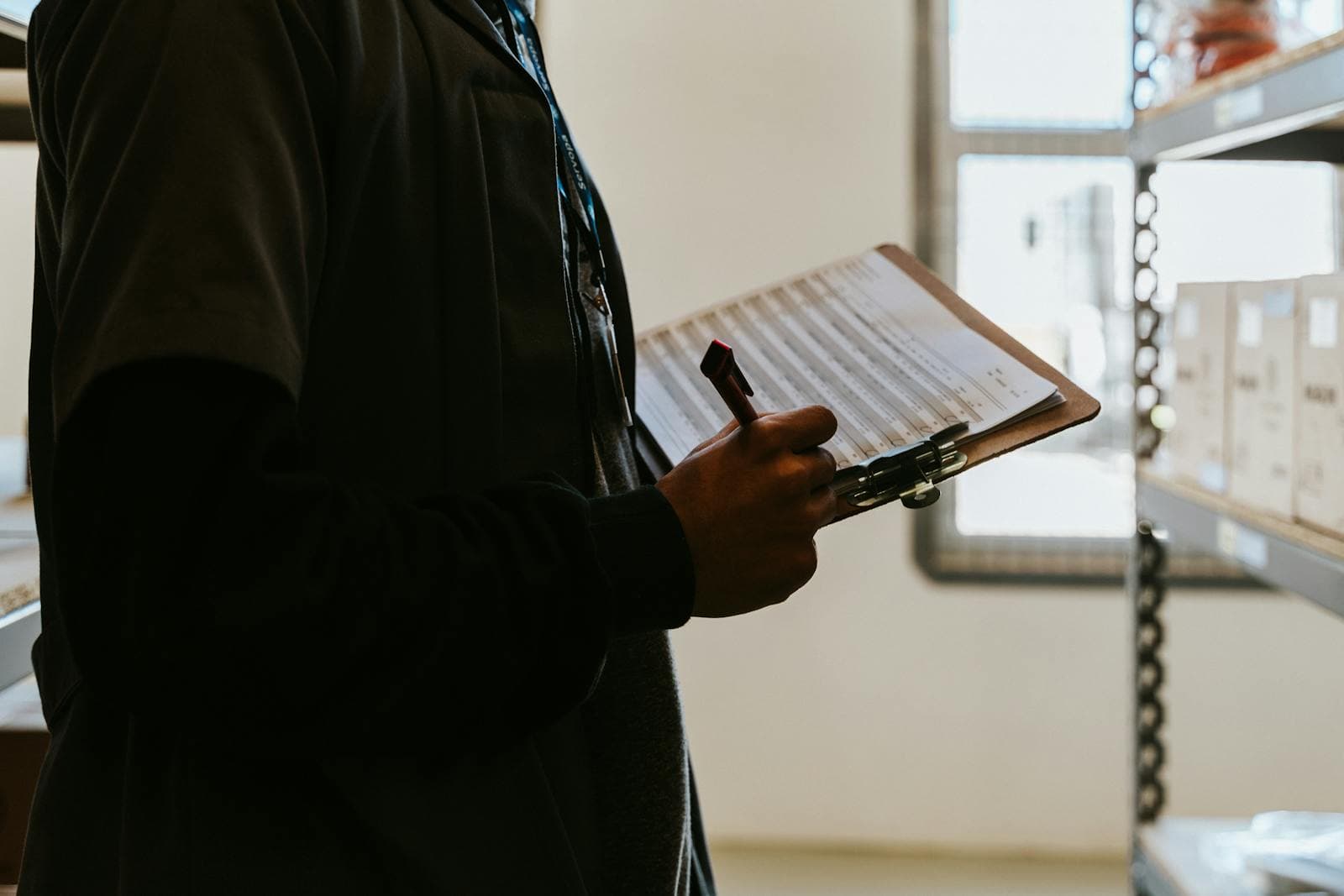 Person med adgangskort fyller ut et sjekklisteskjema på en skriveplate foran lagerhyller.